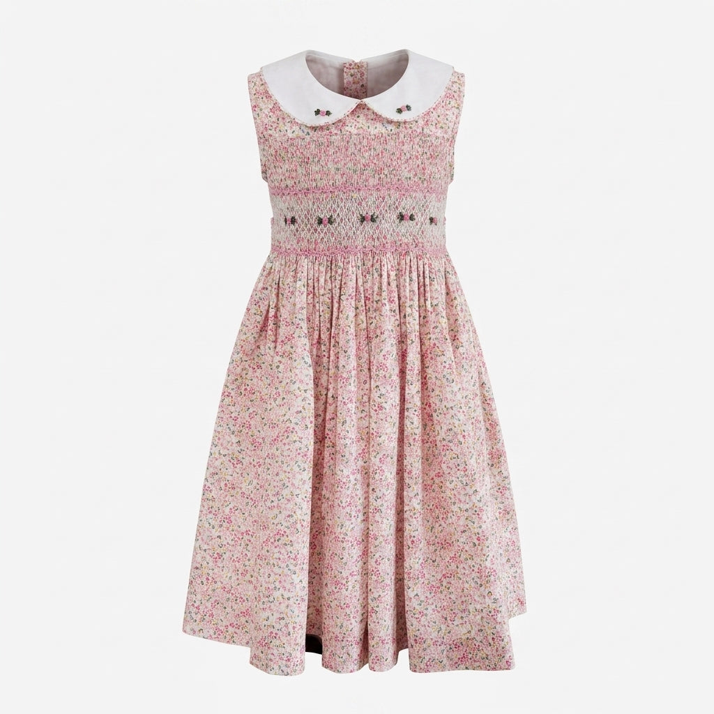 A sleeveless hand smocked dress with a pink and white floral print, featuring a white Peter Pan collar and a smocked bodice with small bow details, shown against a light gray background.