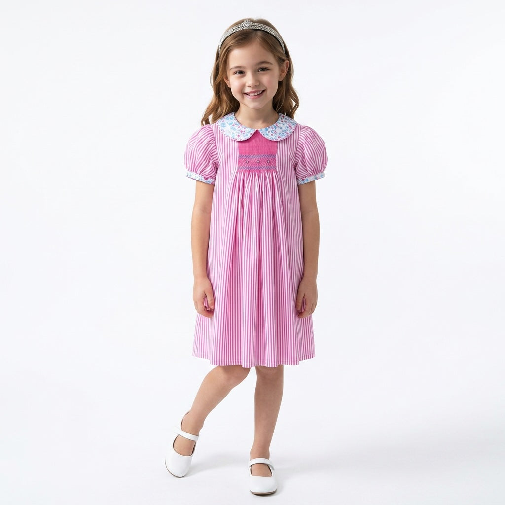 A young girl stands against a studio background wearing a pink and white vertically striped smocked dress with a floral Peter Pan collar.