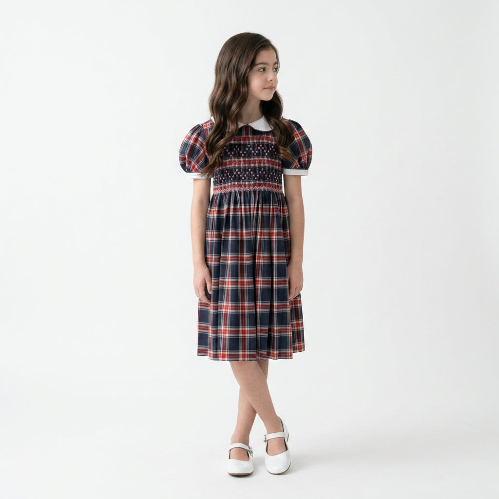 A young girl stands wearing a hand smocked navy plaid dress, a white collar, and puff sleeves.