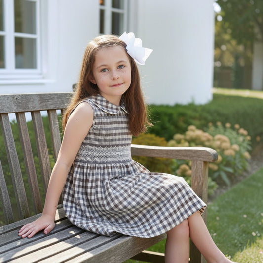Young girl in a checkered dress sitting on a wooden bench outdoors wearing a hand smocked dress.