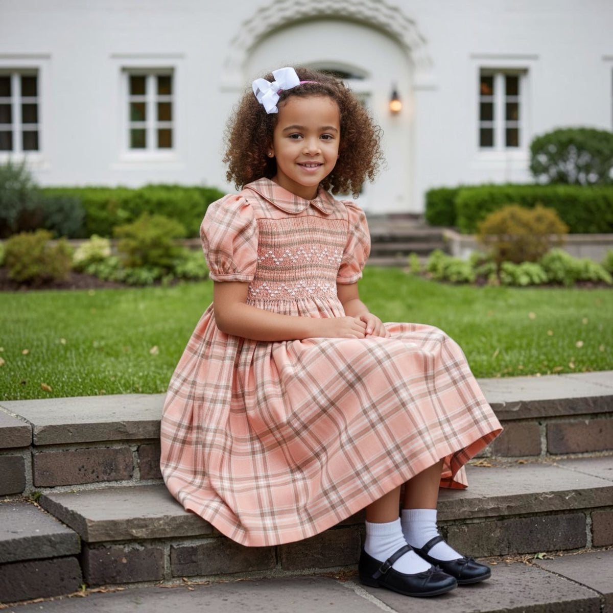 Young girl in an orange plaid hand smocked dress sitting on steps outdoors.