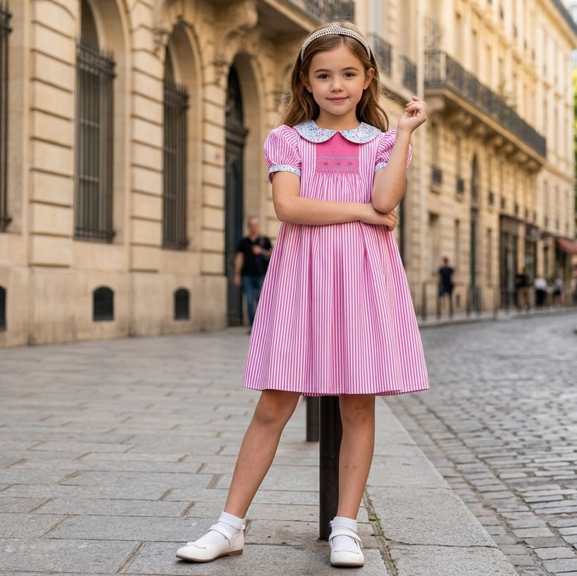 A young girl stands on a cobblestone street  wearing a pink and white vertically striped knee-length hand smocked dress with a floral Peter Pan collar.